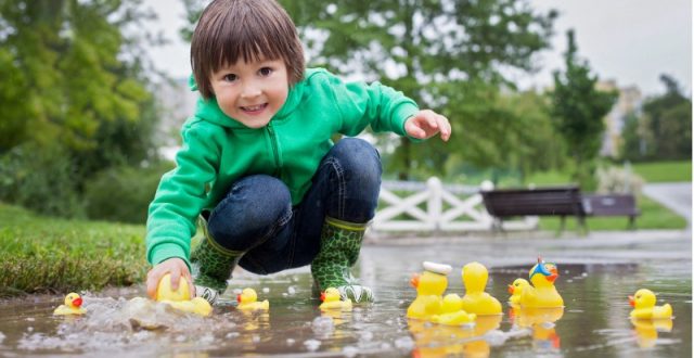 A young boy in a green jacket playing with yellow rubber ducks in a puddle